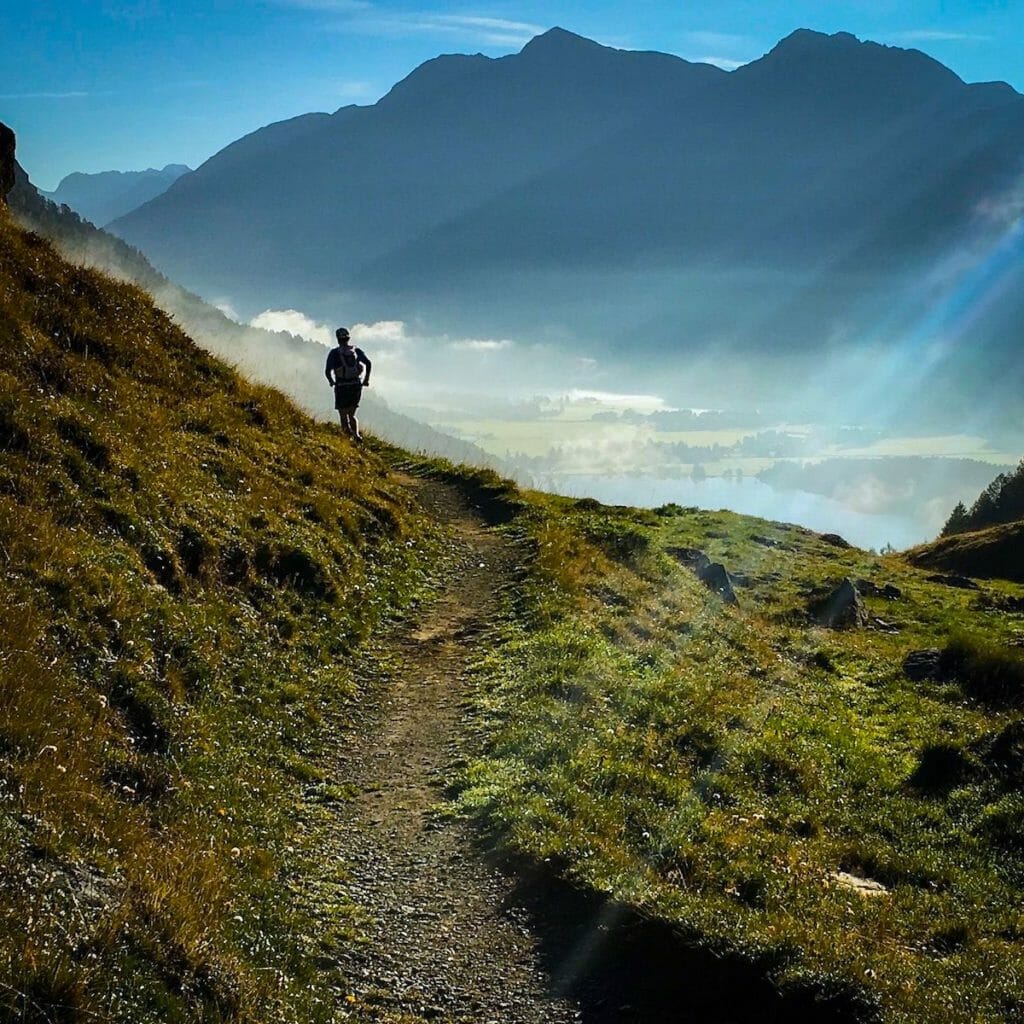 One trailrunner on a single track with mountains and a little lake and sunrays