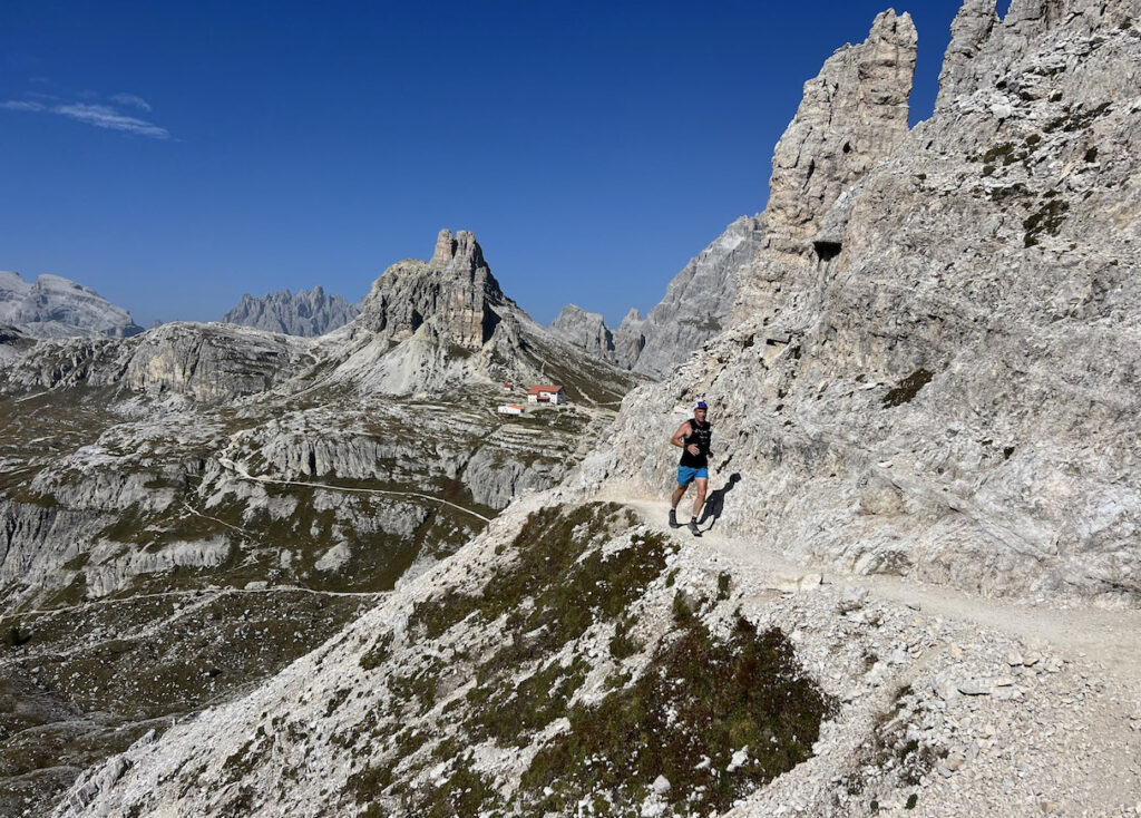 A man trail running in the Dolomites with a mountain hut in the background.
