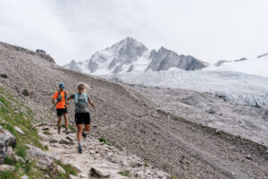 2 runners run down rocky trail with alpine mountains behind